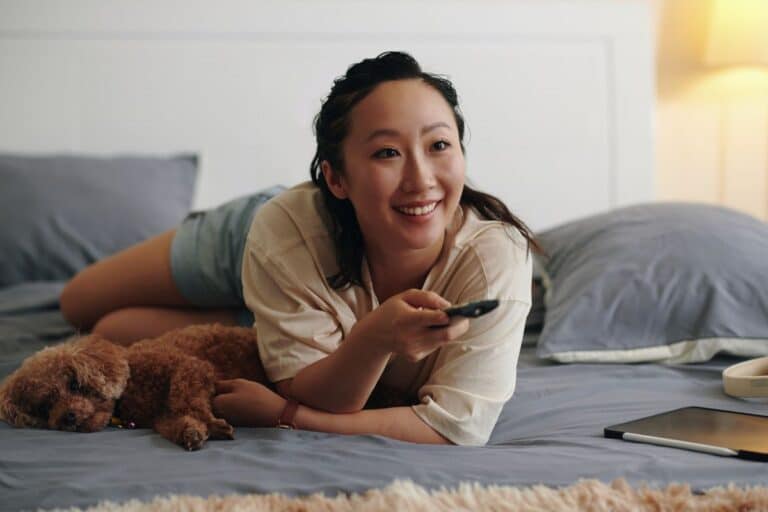 Femme souriante avec chien et télécommande sur lit.