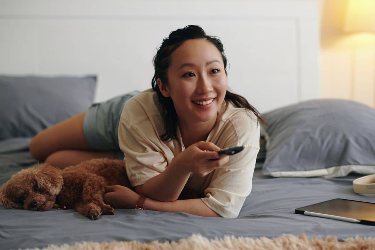 Femme souriante avec chien et télécommande sur lit.