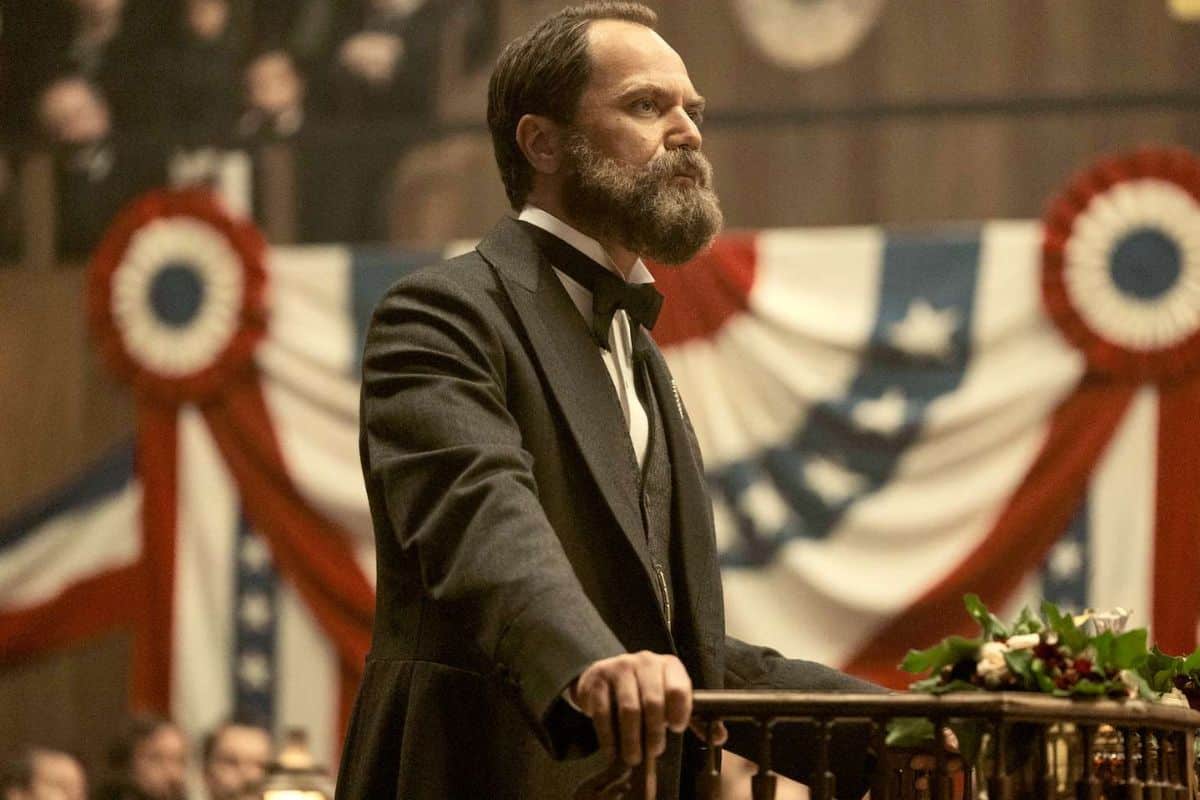 Homme barbu en costume devant des décorations tricolores.