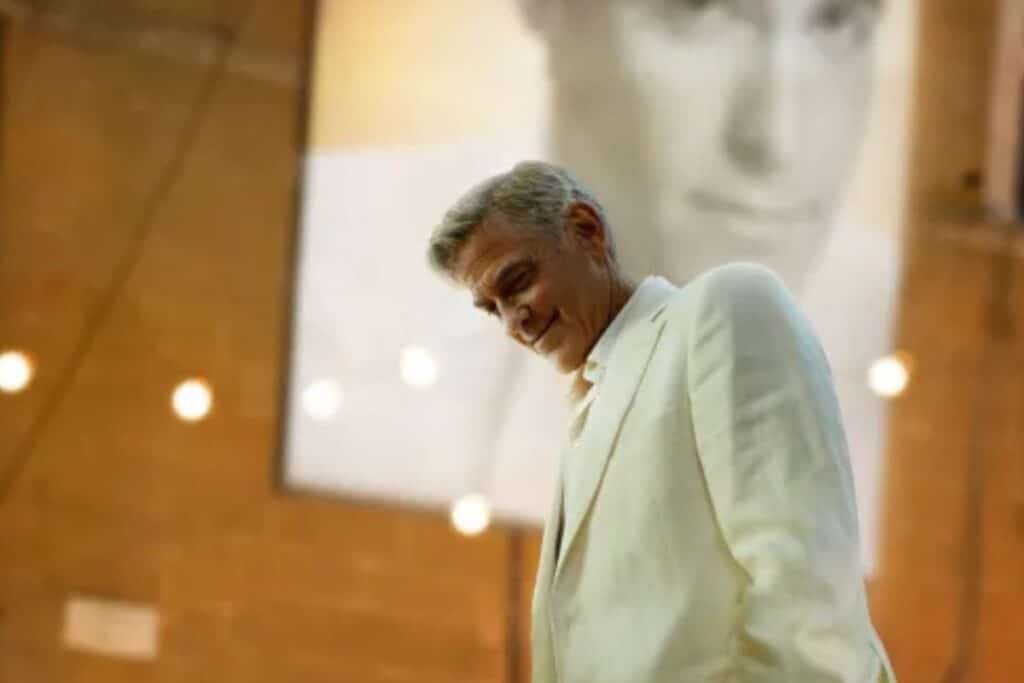 Homme souriant en costume blanc devant une affiche.