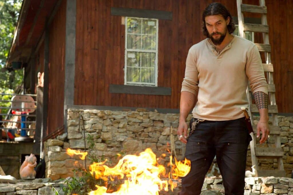 Homme debout devant un feu, maison en arrière-plan.