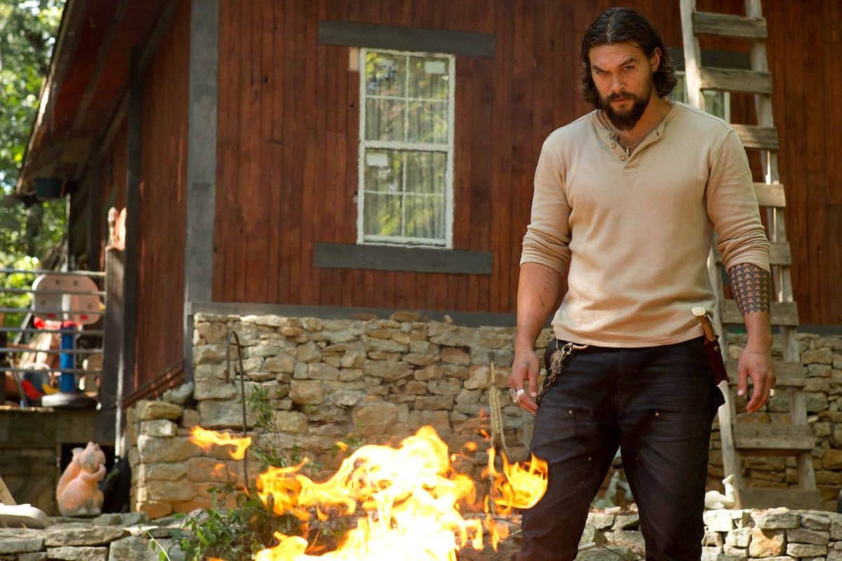 Homme debout devant un feu, maison en arrière-plan.